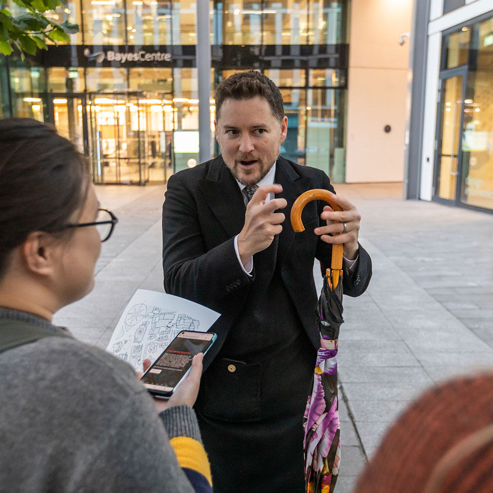 A crowd of onlookers holding their mobile phones and paper maps peer at a costumed performer gesticulating quizzically and holding an umbrella as he speaks to them in front of a backdrop of a glass building sided courtyard lined with paving stones and a tree