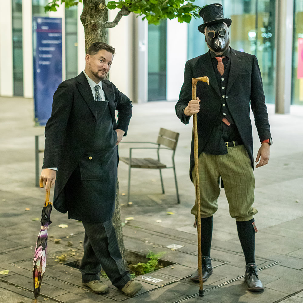 Two figures stand on flagstones in front of a tree, behind them out of focus is a building.  One figure on the left dressed in tails and leaning jauntily on an umbrella is a man staring at the camera smiling, to the right a taller figure with a bird topped wooden staff is dressed with plus fours and an elaborate steampunk style outfit, tartan tie, knee high socks and top hat and stares straight at the camera in a plague doctor mask.