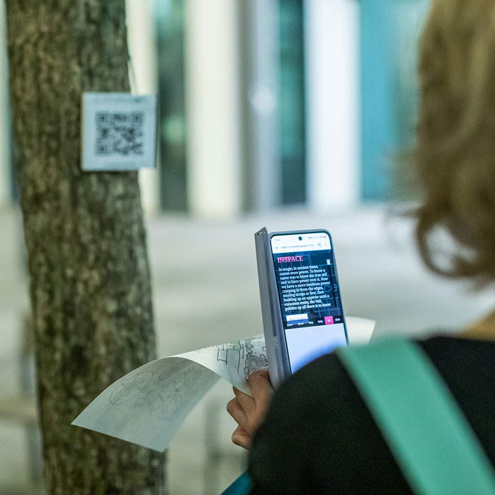 A player stares away from camera and is slightly out of focus, they are pointing they are reading their phone which has game story information, in their had they clutch a hand drawn map, in the back of shot is an out of focus tree