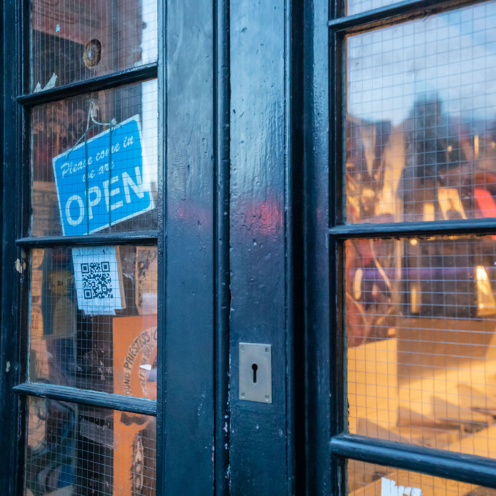 A greeny blue door, shot askance occupies the picture frame, lit from within the golden light shines through the glass panes on the door, inside shelves of books and comics can be glimpsed.  A prominent keyhole is centre shot on the door as is a sig saying ‘Pleas come in we are open’ and a QR code hanging from it.