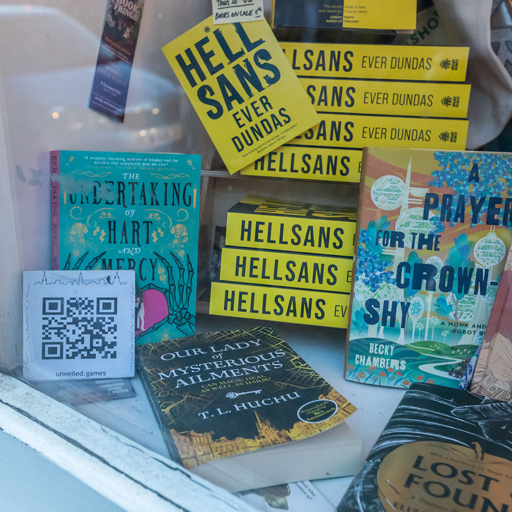 A shot through the glass of a bookshop window, colourful books displayed artfully, mostly yellow and greens, A QR code and sign saying ‘unveiled.games’ with an Edinburgh skyline artwork visible, perched atop it a stylised rook.