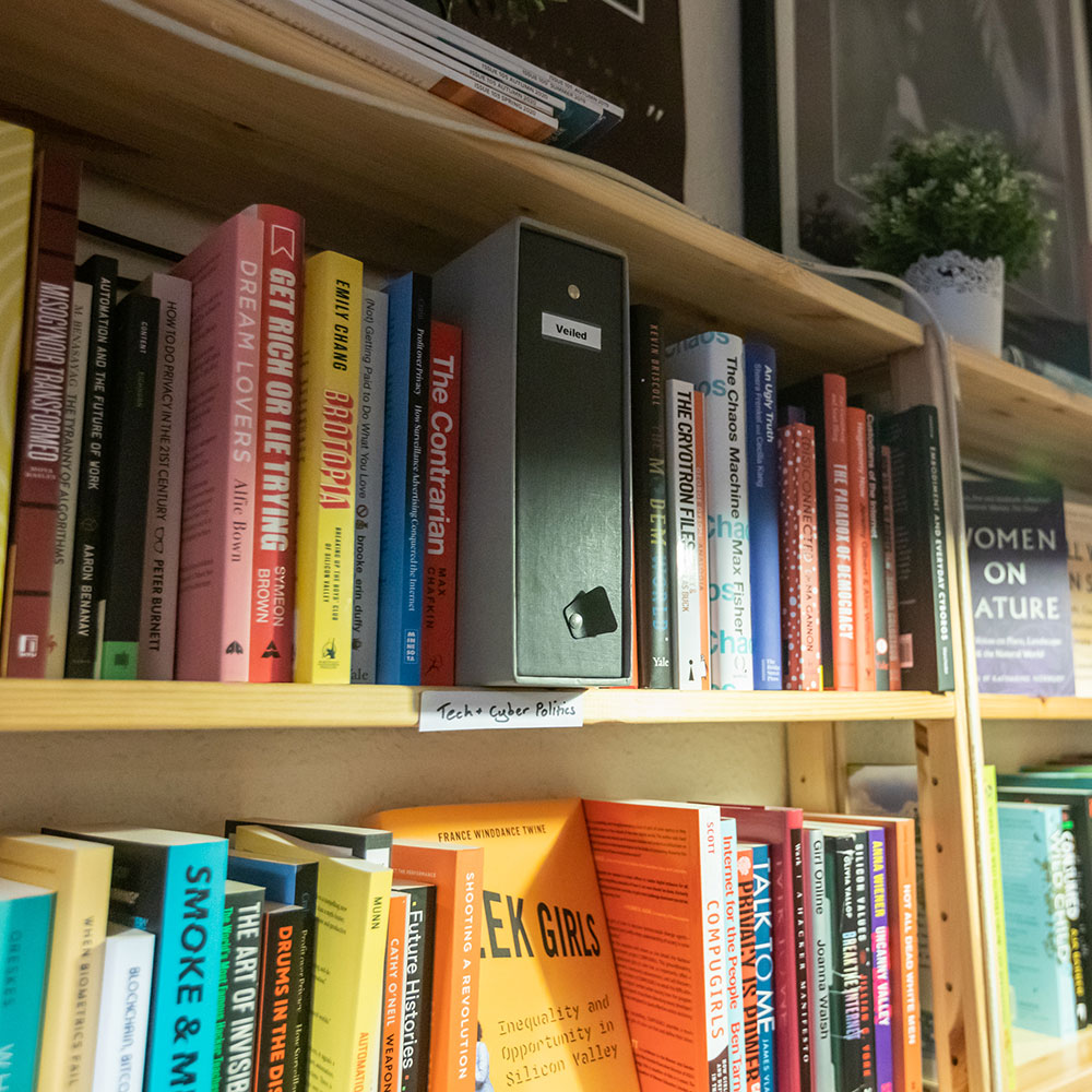 A pale wooden bookshelf is visible stretching away to the right of shot, a plant perched atop it.  A handwritten paper sign states ‘Tech & Cyber Politics.’ And the two shelves visibile are lined with related books.  Centre shot is a mysterious grey folder that says ‘Veiled’ on it’s spine.